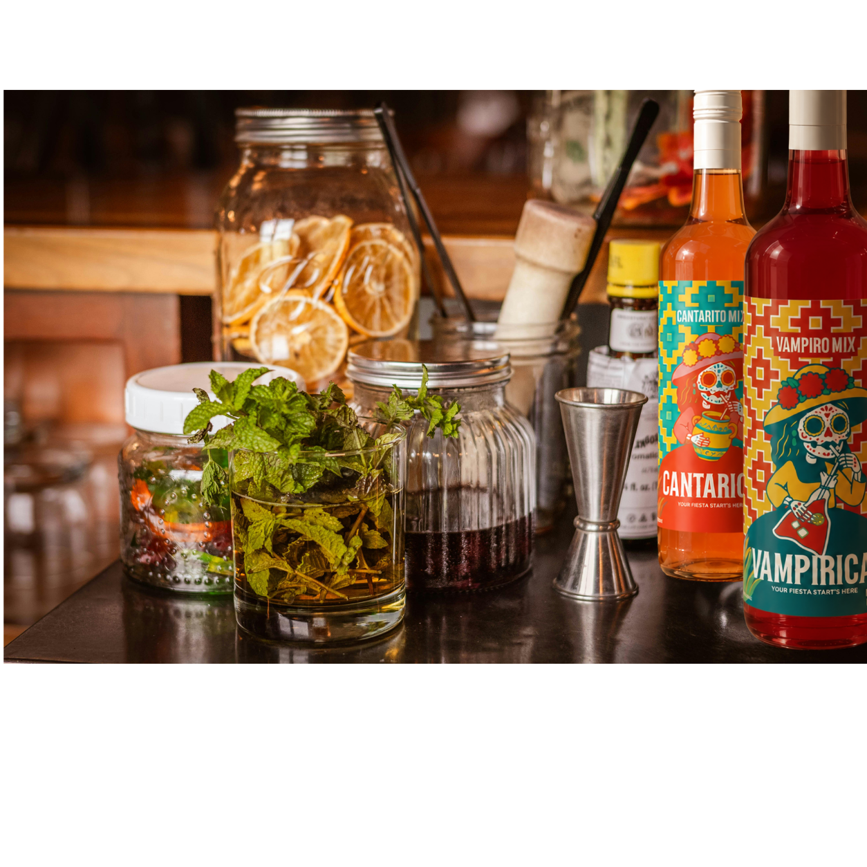 Both signature mixer bottles on a bar with mint, citrus jars and a jigger
